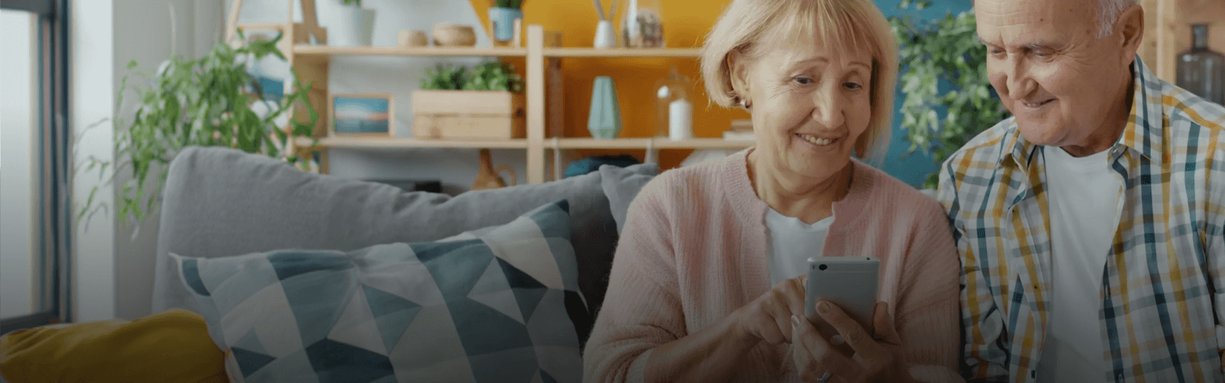 A smiling elderly couple sits on a couch, looking at a smartphone held by the woman.