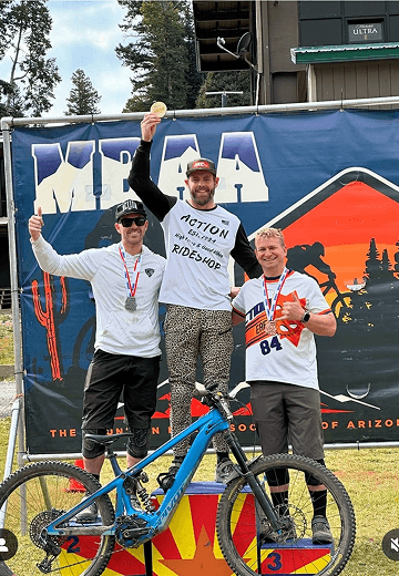 Three male mountain bikers celebrate on a podium with their medals. The first-place winner holds up a gold medal, flanked by second and third-place winners with silver and bronze medals, respectively. A blue mountain bike rests in front.