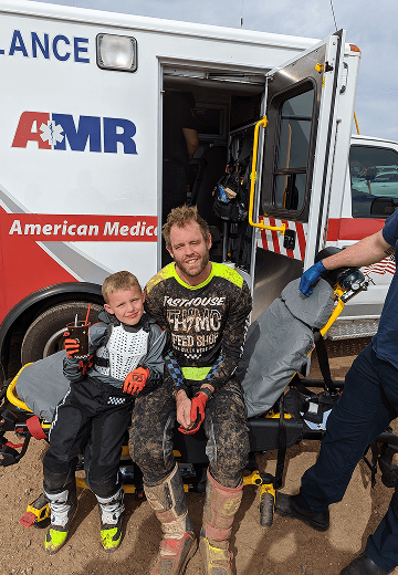 A mud-covered man and young boy in motocross gear smile while sitting on a stretcher in front of an open AMR ambulance.