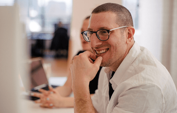 A smiling man with glasses and a white shirt looks right, his fingers touching his lower lip, in a bright, blurred office background.