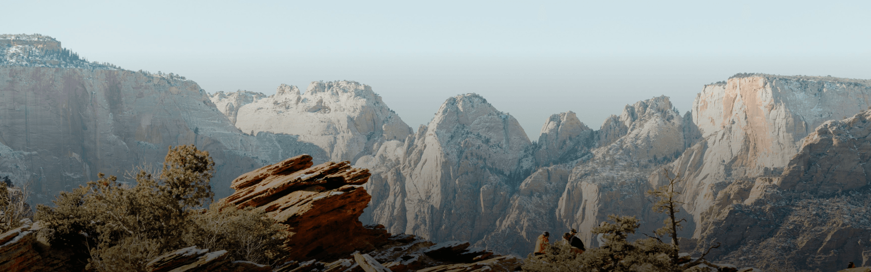 Expansive view of a majestic mountain range with red and tan sandstone cliffs, dotted with snow and evergreen trees, seen from a high overlook where a few people stand.