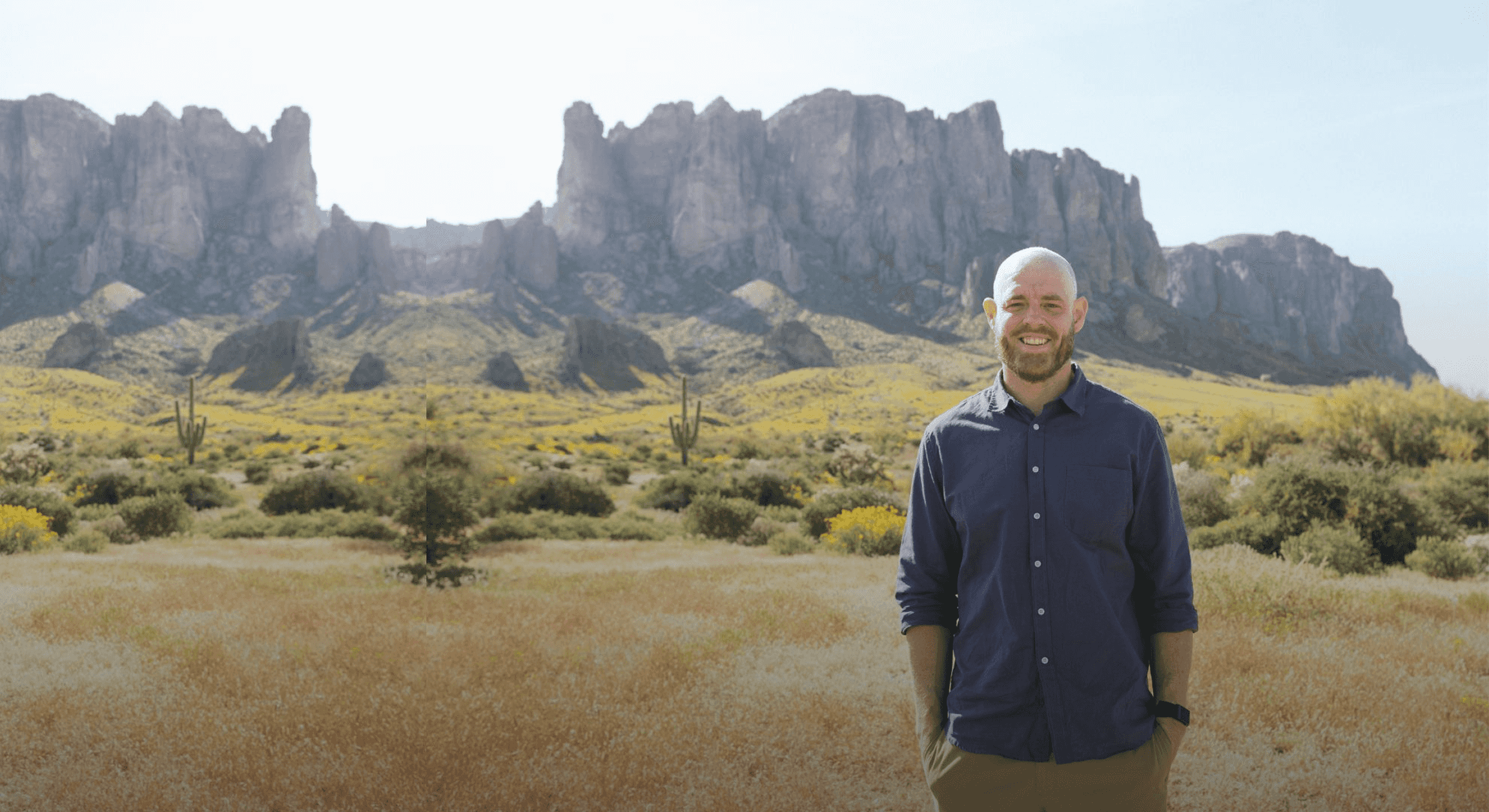 Smiling man with a beard stands in a desert landscape featuring large mountains, saguaro cacti, and yellow wildflowers.