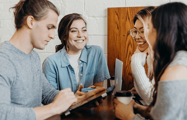 Four smiling young adults are gathered around a table with a tablet and coffee cups, engaging in conversation or collaboration.