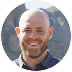 Smiling man with a bald head and a beard in a blue shirt, standing outdoors with mountains in the background.